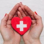La imagen muestra unas manos con las uñas pintadas de rojo mostrando un corazón con una cruz blanca en medio. Accidente laboral que es y que seguro lo cubre
