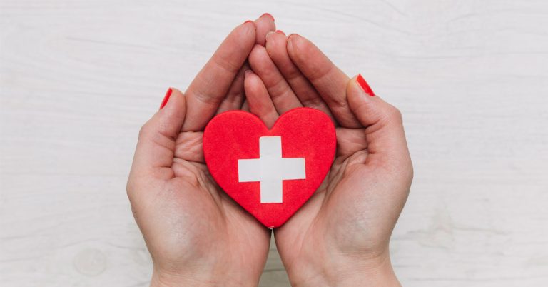 La imagen muestra unas manos con las uñas pintadas de rojo mostrando un corazón con una cruz blanca en medio. Accidente laboral que es y que seguro lo cubre